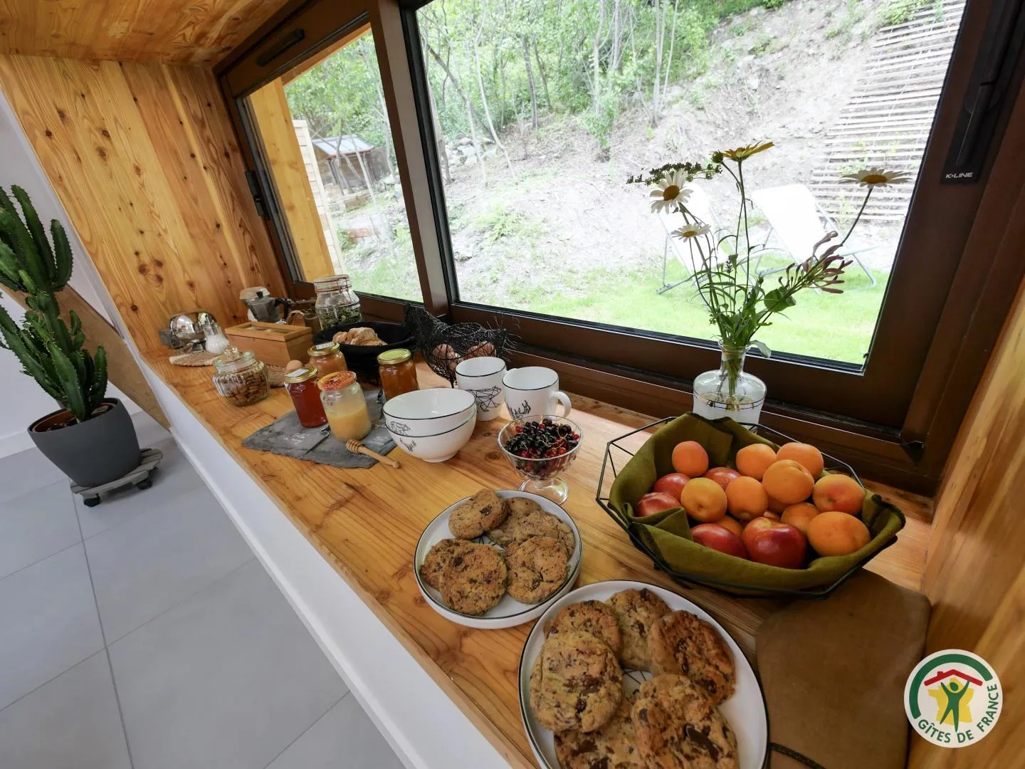 Une table dressée devant la fenêtre, avec confitures maison, fruits frais et cookies